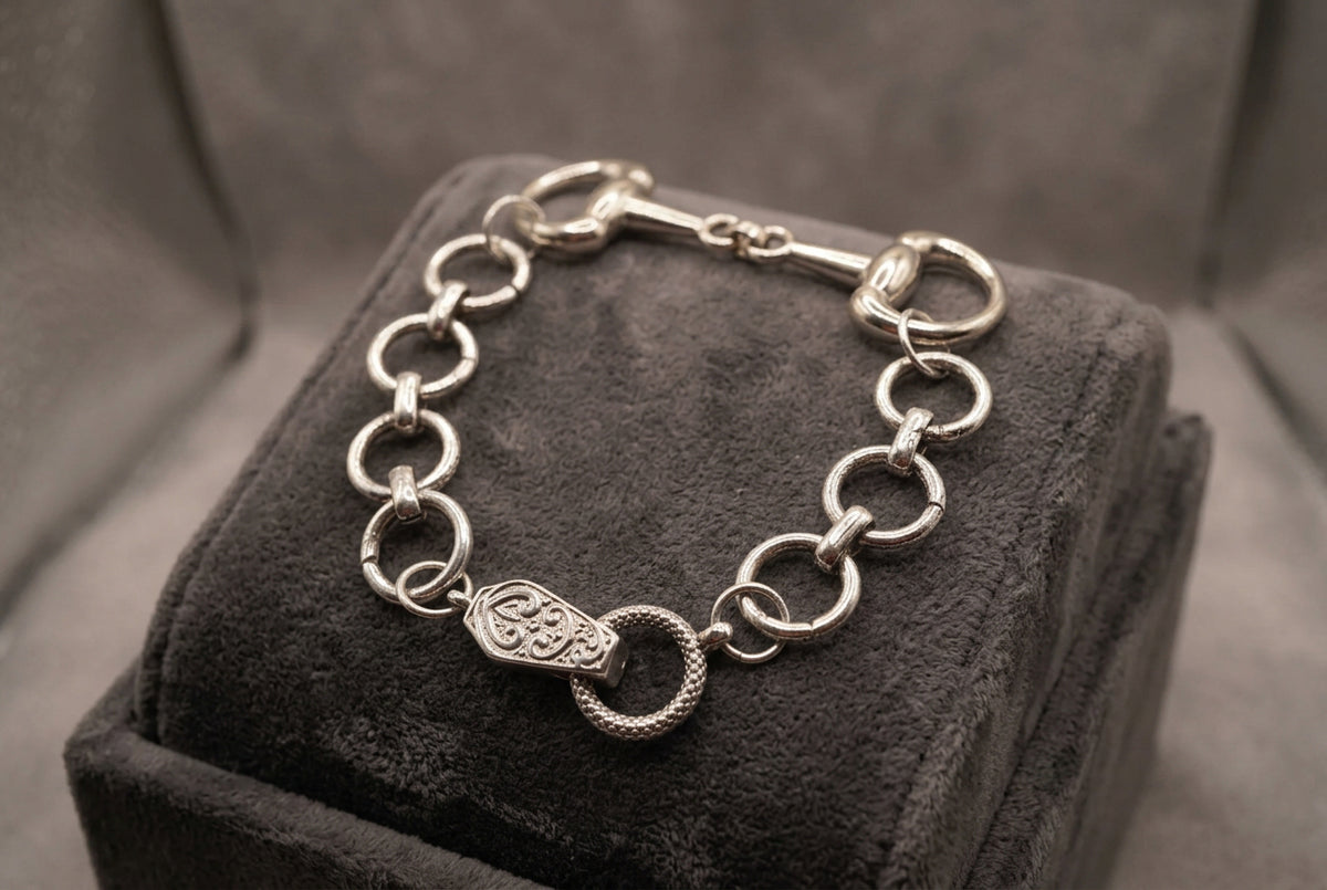 A silver-tone chain bracelet with a horsebit clasp and a central engraved charm, displayed on a neutral gray background.
