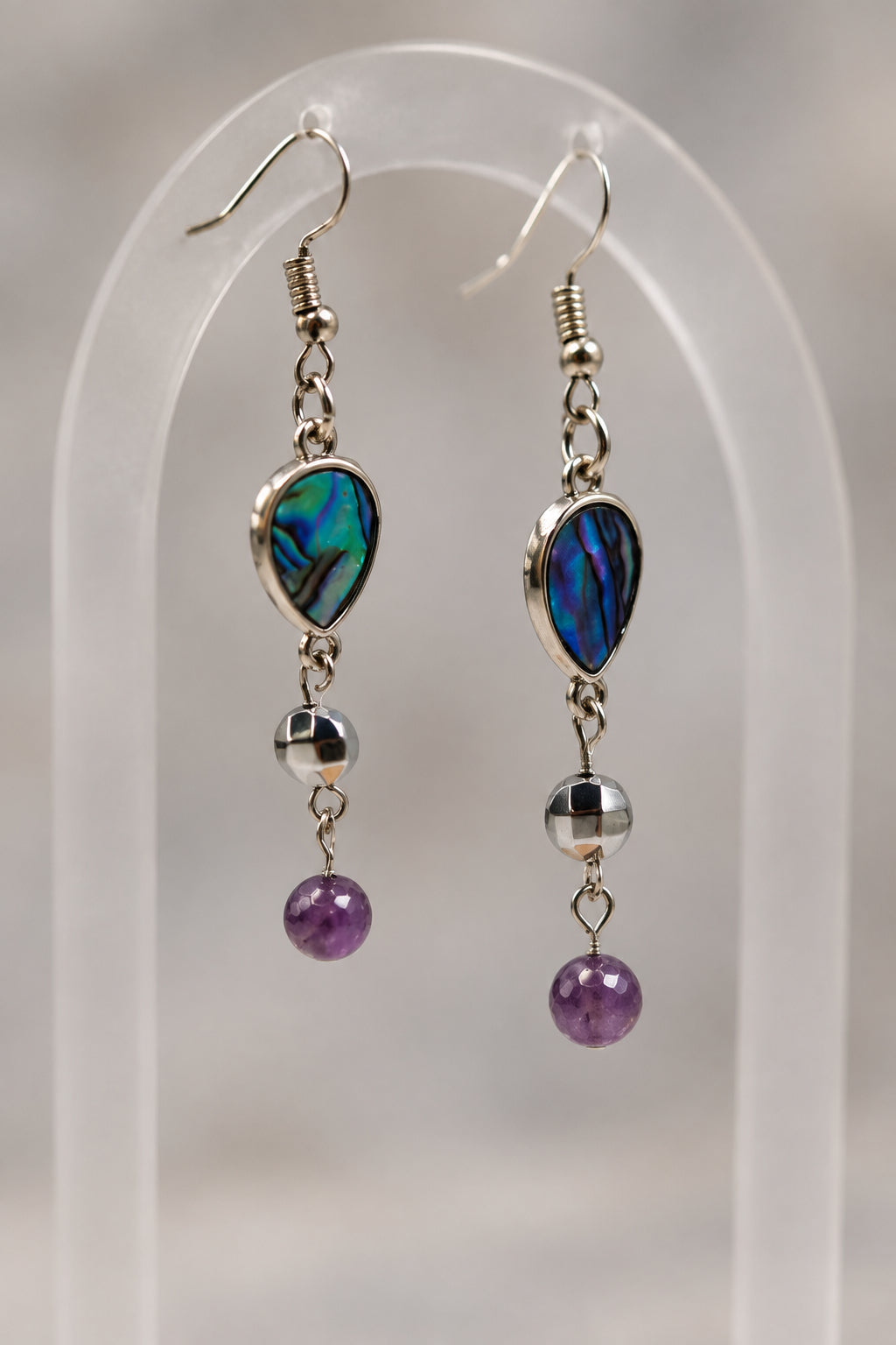 A pair of silver dangle earrings with iridescent green and blue teardrop beads, small silver beads, and dark purple round beads, displayed on a clear acrylic stand against a light gray background.