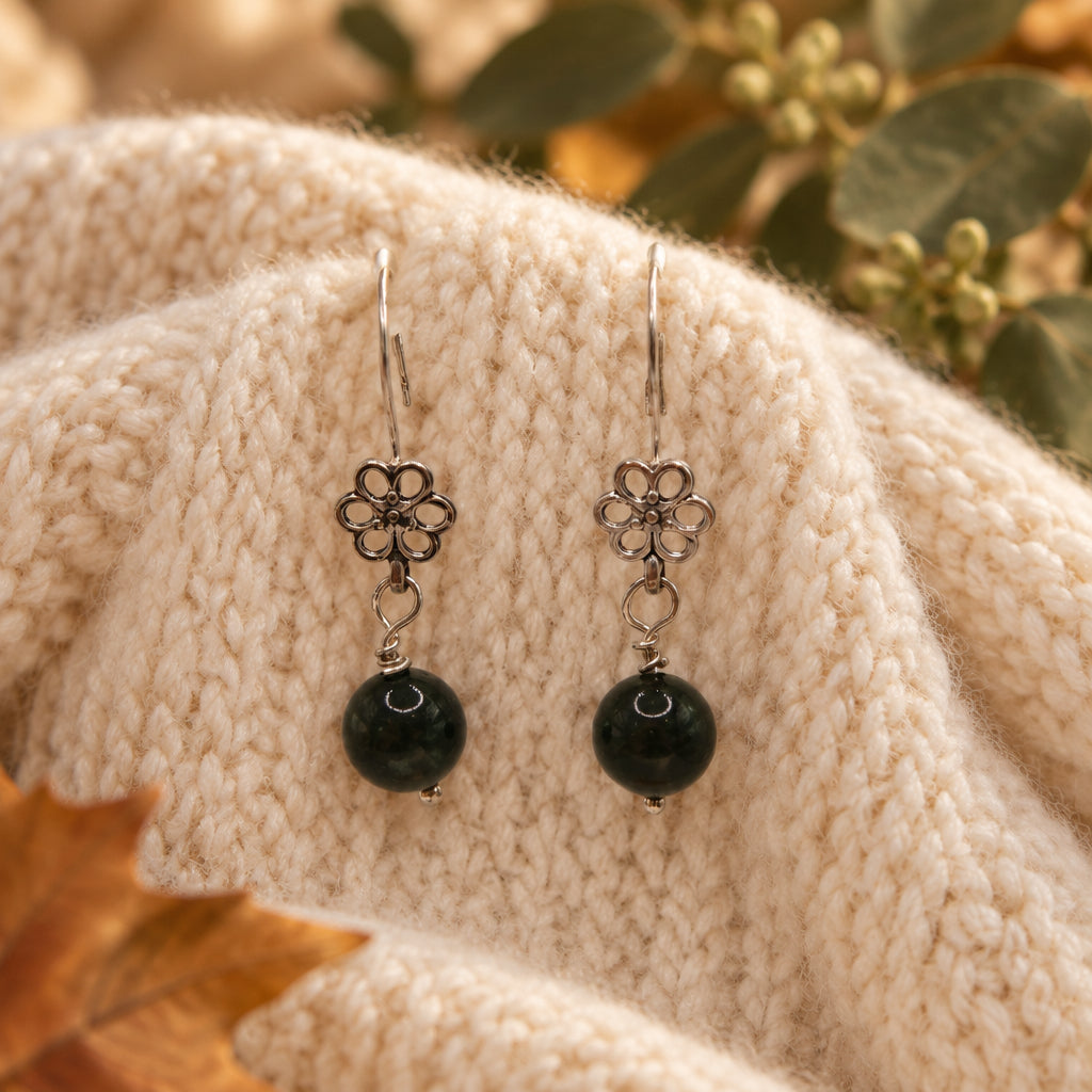 A pair of dangling earrings with silver-tone flower-shaped charms and round black beads, displayed on a clear acrylic stand against a light gray background.