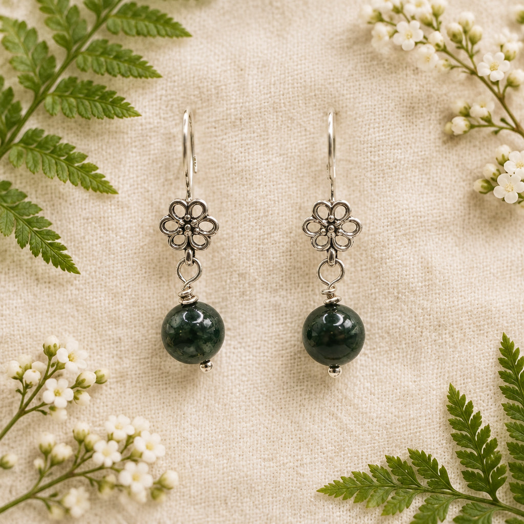 A pair of dangling earrings with silver-tone metal flower charms and round black beads, displayed on a clear acrylic stand against a light gray background.