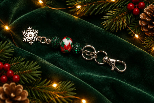 A silver-tone metal keychain charm featuring a small silver snowflake, dark green and red patterned beads, and a lobster clasp, shown on a plain gray background.