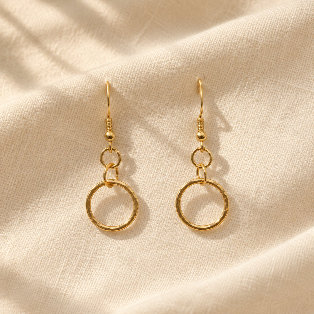 A pair of gold dangle earrings with a large open circular ring hanging from a small ring and beaded chain, displayed on a clear stand against a light gray background.