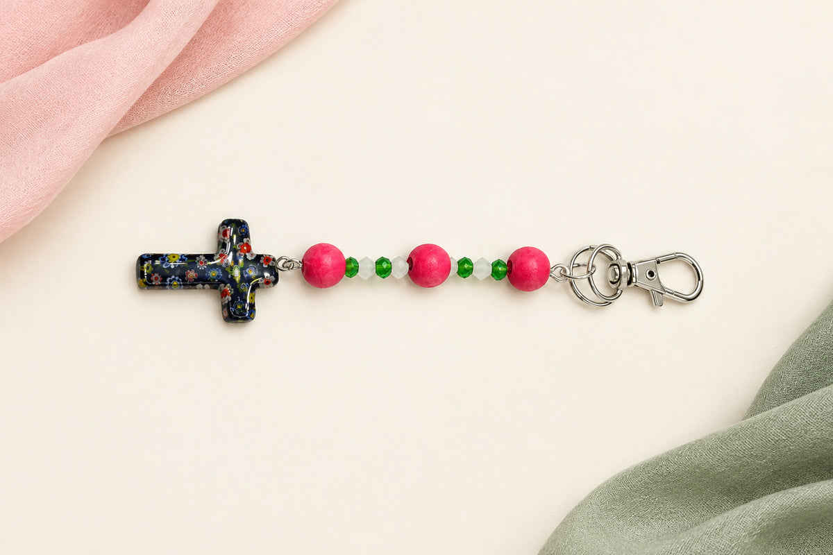 A beaded rosary keychain featuring a small metallic cross, pink wooden beads, green spacer beads, and silver metal key rings, displayed on a plain light gray background.