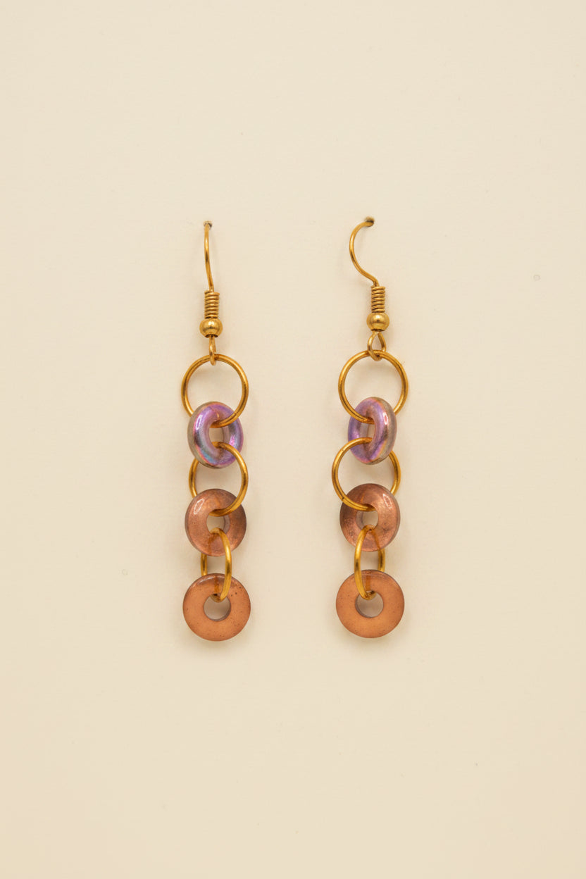 A pair of handmade dangle earrings with metallic copper-colored rings and iridescent purple accents, displayed on a white 'River Inspired' backing card against a neutral gray background.