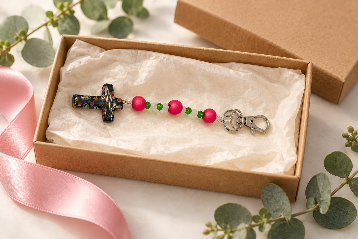 A rosary-style keychain with a small metallic cross, pink and green beads, and a silver lobster clasp on a plain light gray background.