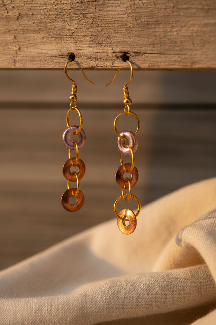 A pair of handmade dangle earrings with copper-colored circular links and iridescent pink and purple beads, displayed on a white card labeled 'River Inspired Handcrafted Jewelry' against a neutral gray background.