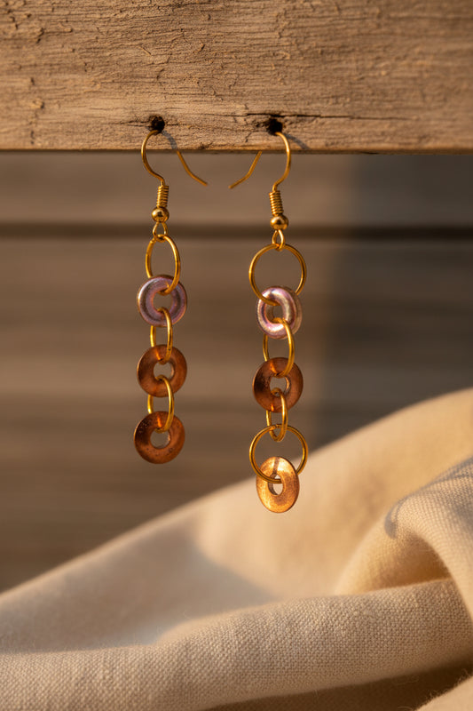 A pair of handmade dangle earrings with copper-colored circular links and iridescent pink and purple beads, displayed on a white card labeled 'River Inspired Handcrafted Jewelry' against a neutral gray background.