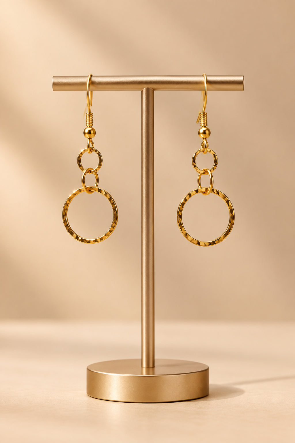A pair of gold dangle earrings with simple circular hoop charms, displayed on a clear plastic stand against a light gray background.
