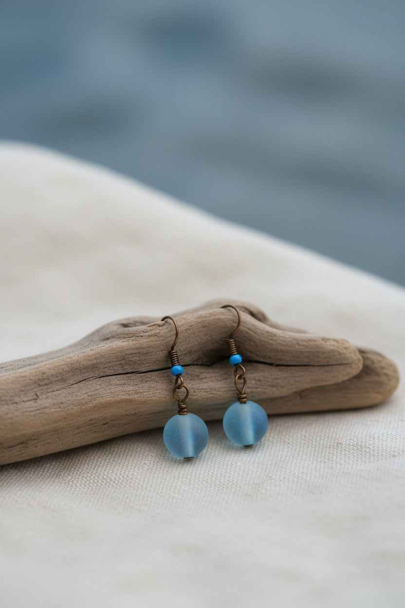 A pair of handmade dangle earrings with iridescent blue beads and small blue beads on gold-toned hooks, displayed on a white card labeled River Inspired.
