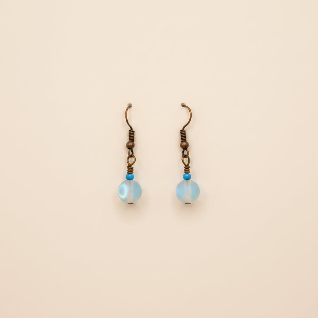 A pair of handmade dangle earrings with iridescent blue beads and bronze hooks, displayed on a white 'River Inspired' backing card against a neutral gray background.
