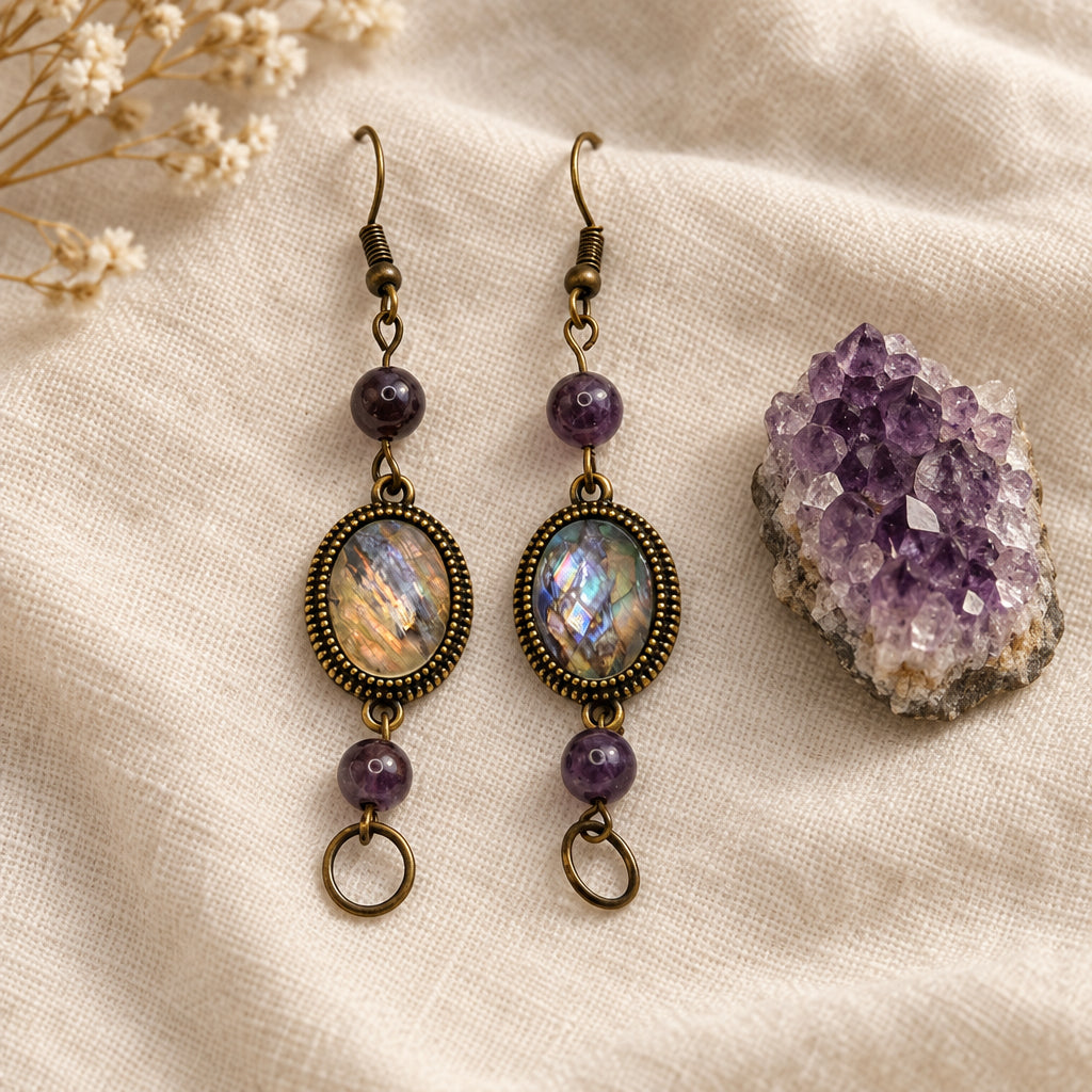 A pair of handmade dangle earrings displayed on a white card labeled 'River Inspired Handcrafted Jewelry.' The earrings have oval iridescent shell-like centers in an antique bronze-toned frame, purple beads, and small dangling rings, photographed against a plain light background.