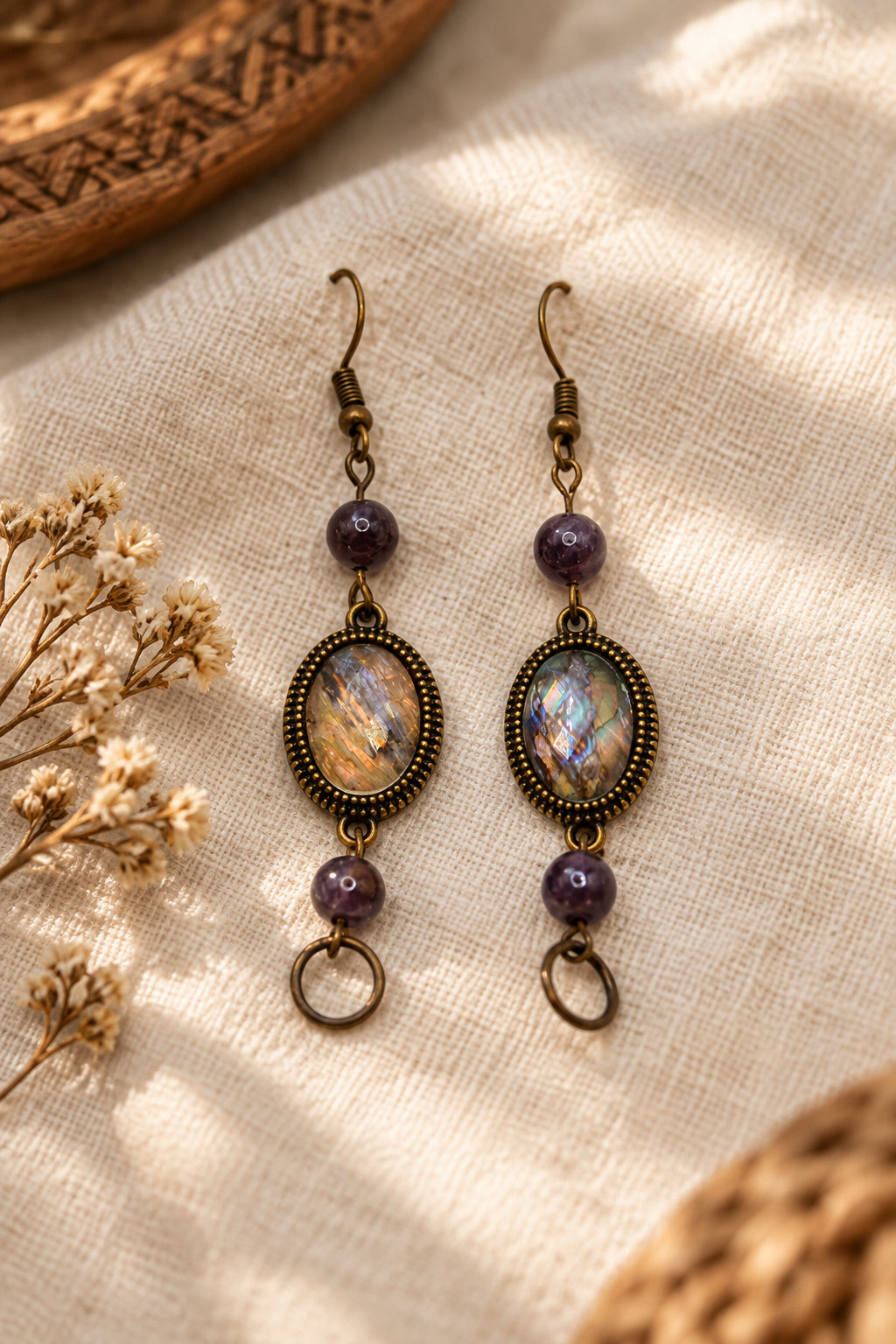 A pair of handmade dangle earrings displayed on a white backing card. The earrings feature oval iridescent cabochon stones with an antique bronze-toned metal frame, purple beads, and small dangling rings at the bottom.