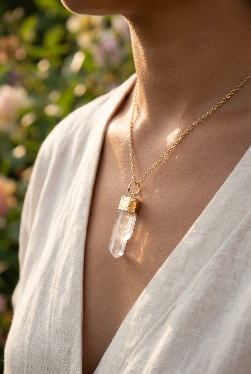 A clear quartz crystal pendant necklace with a gold cap, displayed on a black velvet necklace bust with a hand holding the left side.