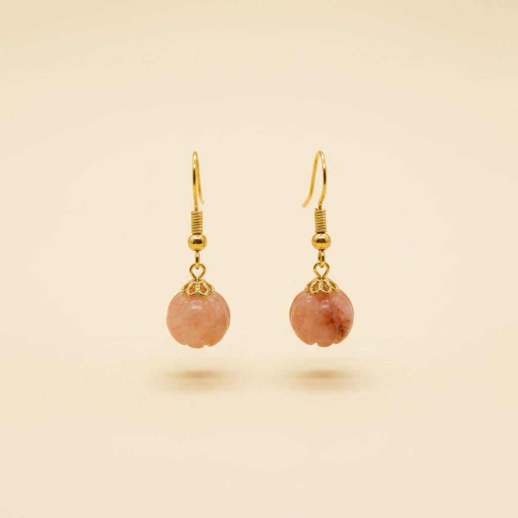 A pair of gold-tone dangle earrings with peach-pink flower-shaped charms, displayed on a clear acrylic stand against a neutral gray background.