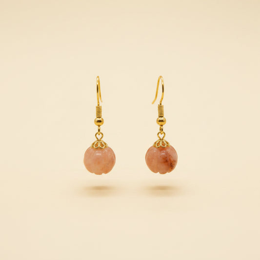 A pair of gold-tone dangle earrings with peach-pink flower-shaped charms, displayed on a clear acrylic stand against a neutral gray background.