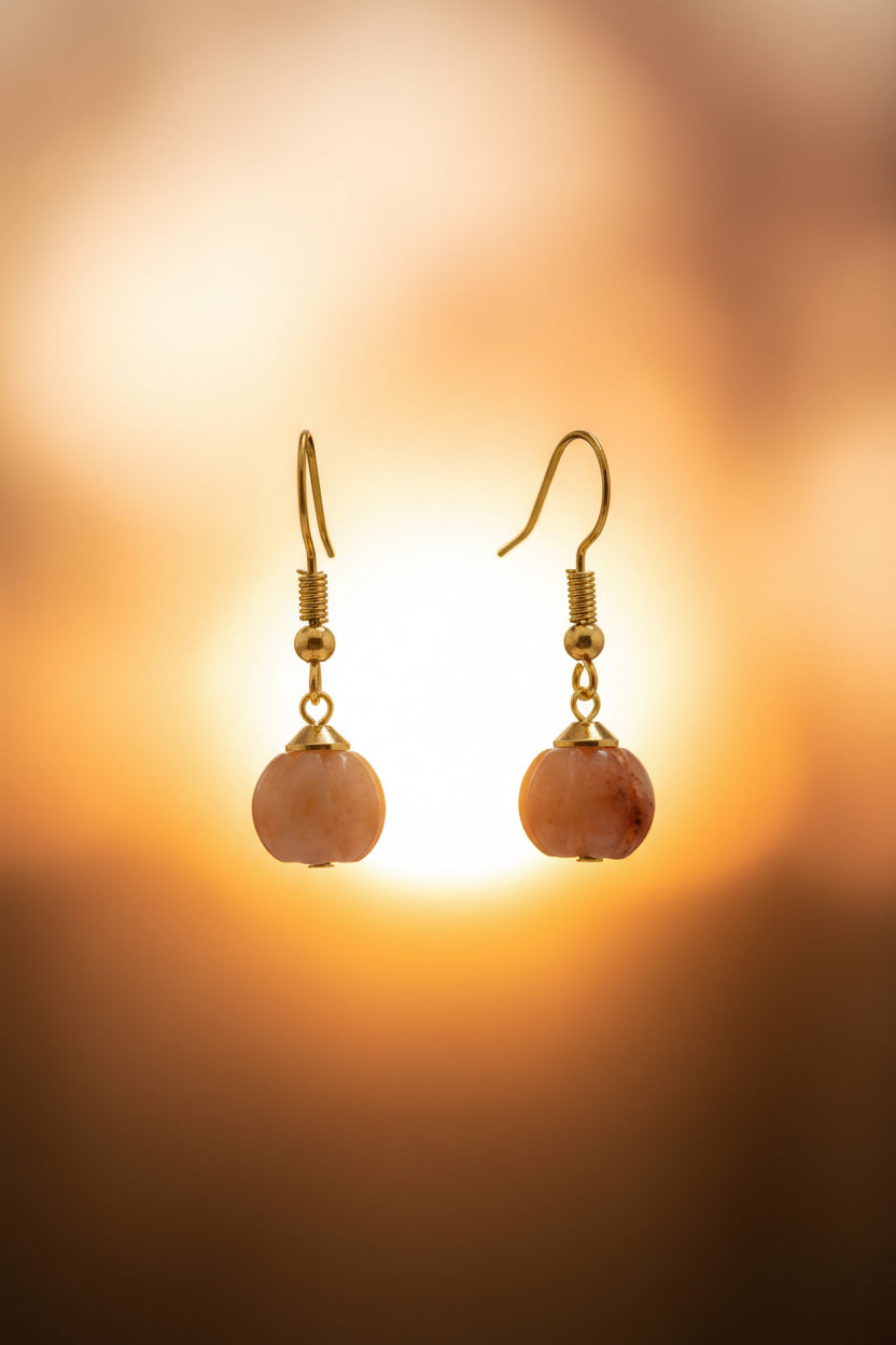 A pair of gold-tone dangle earrings with peach-pink stone drops, displayed on a clear acrylic stand against a light gray background.