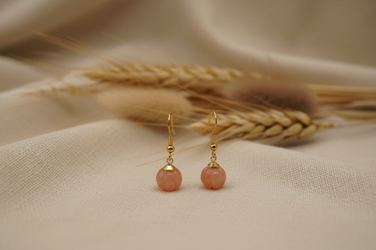 A pair of gold-tone dangle earrings with round, peach-pink stone pendants, displayed on a clear acrylic stand against a neutral gray background.