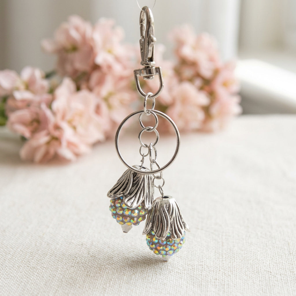 A silver-tone metal keychain featuring two dangling flower-shaped charms embellished with iridescent rhinestones, hanging from a clear plastic display stand.