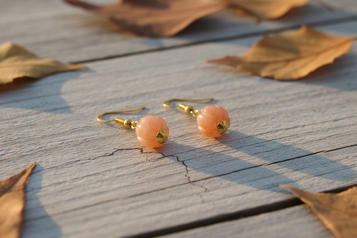 A pair of gold-tone dangle earrings with peach-pink stone pendants, displayed on a clear acrylic stand against a light gray background.