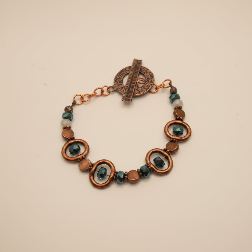A handmade beaded bracelet with copper-toned metal rings, faceted brown beads, and dark teal-green glass beads, featuring a rustic copper toggle clasp, shown on a plain light gray background.