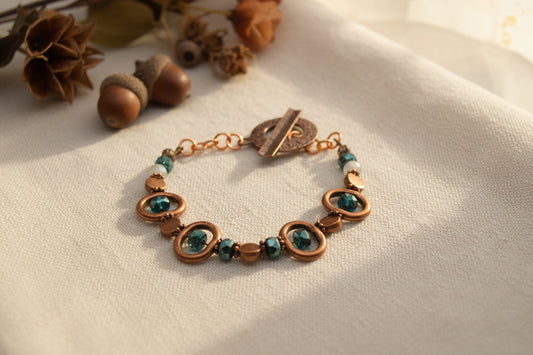 A handmade beaded bracelet with copper-toned metal rings, faceted brown beads, and dark green iridescent beads, featuring a rustic copper toggle clasp, shown on a plain light gray background.