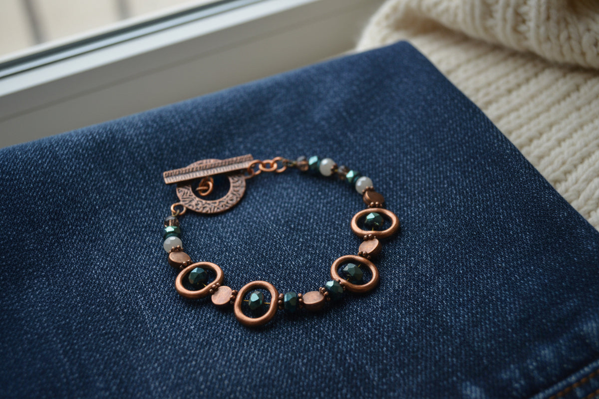 A handmade beaded bracelet with copper-toned metal links, faceted brown beads, and dark green glass beads, featuring a rustic copper toggle clasp, shown on a plain light gray background.