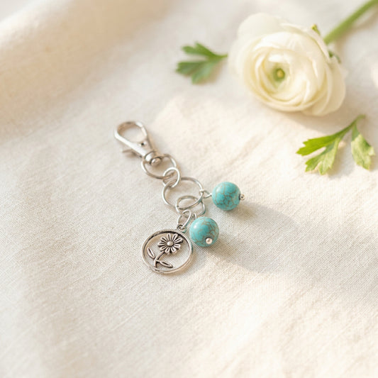 A silver-toned keychain charm featuring a tree of life medallion and two small turquoise beads, shown on a plain light gray surface.
