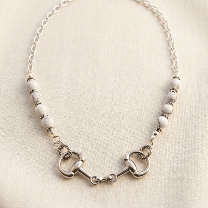 A silver-toned beaded necklace featuring a horse bit charm in the center, laid flat on a white surface.