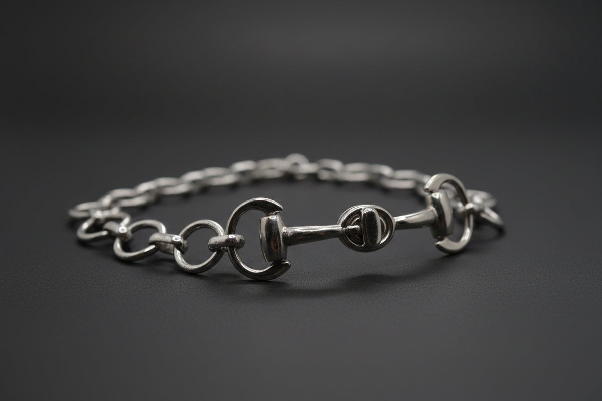 A silver horse bit chain bracelet with interlocking rings and a central horse bit clasp, displayed on a neutral light gray background.