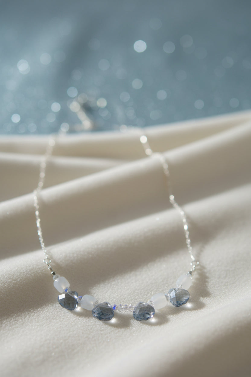 A delicate silver-toned chain necklace featuring a row of faceted light blue beads and clear round spacer beads, displayed against a plain light gray background.