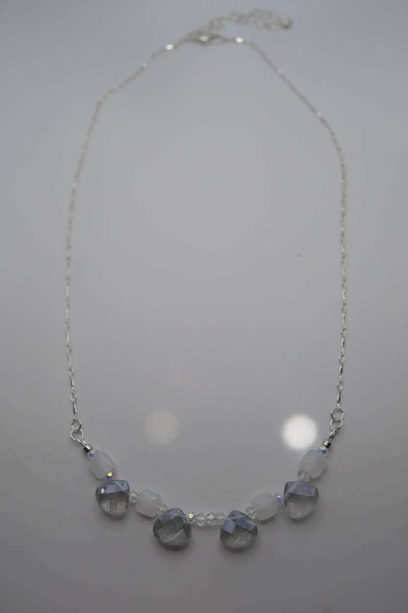 A delicate silver chain necklace featuring a row of faceted clear beads and three larger round light blue beads at the center, displayed against a plain light gray background.
