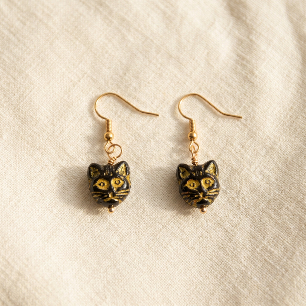 A pair of dangling earrings with small black cat-shaped charms decorated with gold and yellow details, hanging from gold fishhook wires on a plain light gray background.