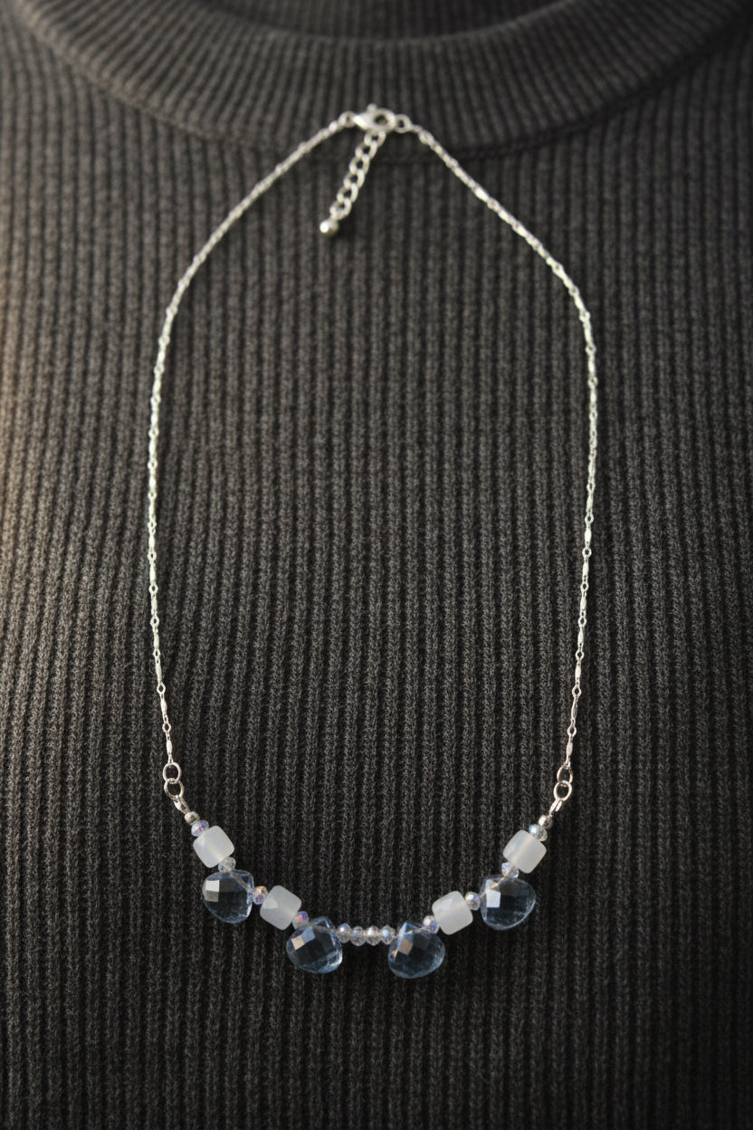 A delicate silver chain necklace featuring a row of faceted light blue beads and clear crystal accents, displayed against a plain light gray background.
