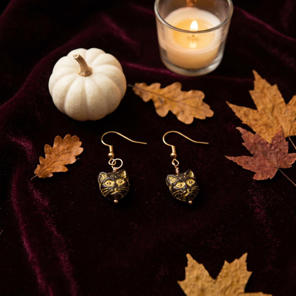 A pair of dangling earrings shaped like black cat faces with yellow eyes and whiskers, hanging from gold-tone hooks on a plain light gray background.