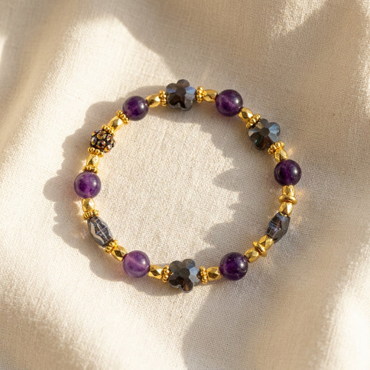 A beaded stretch bracelet featuring purple round beads, gray faceted beads, and shiny gold spacer beads, arranged in a circle on a plain light gray background.