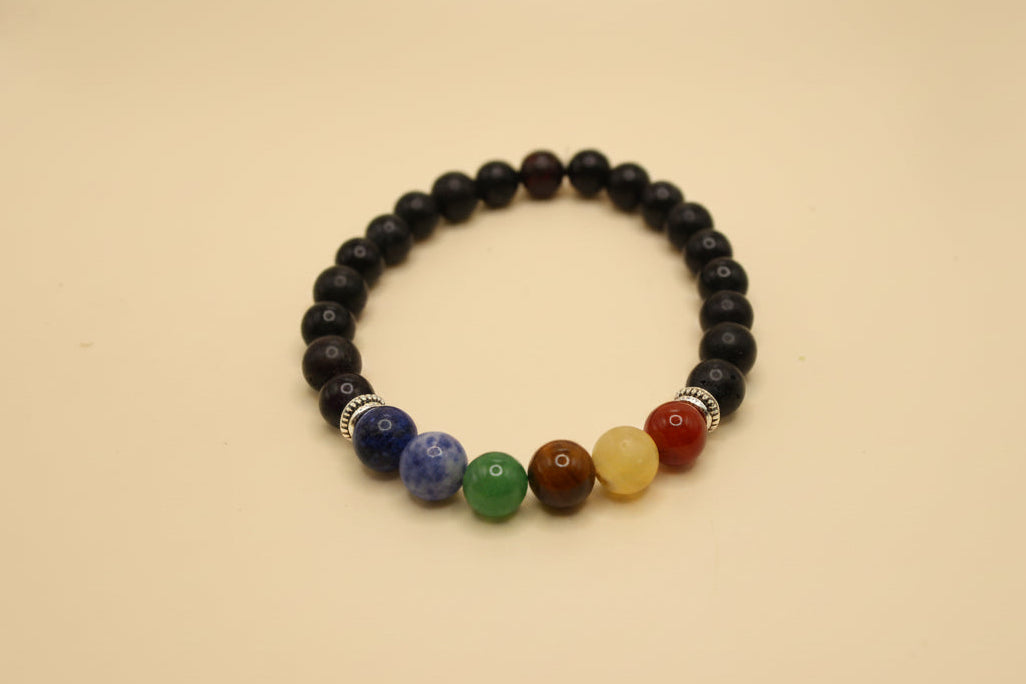 A beaded stretch bracelet featuring a majority of glossy black beads, with a front section of seven colorful round beads representing the chakras in blue, purple, green, brown, yellow, orange, and red, separated by small silver spacer beads, displayed on a light gray background.