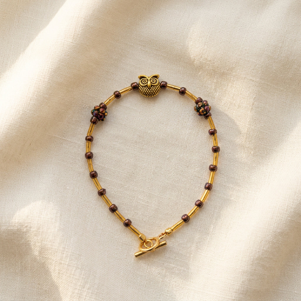 A gold beaded bracelet with dark brown floral accents and a small gold charm, displayed on a plain light gray background.