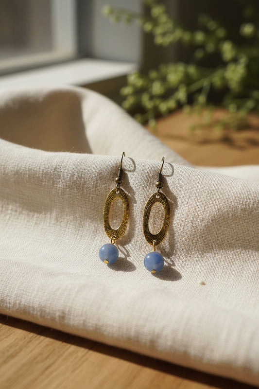 A pair of dangling earrings displayed on a white card. Each earring has a textured gold oval frame with a small blue bead hanging below and dark metal hooks at the top.
