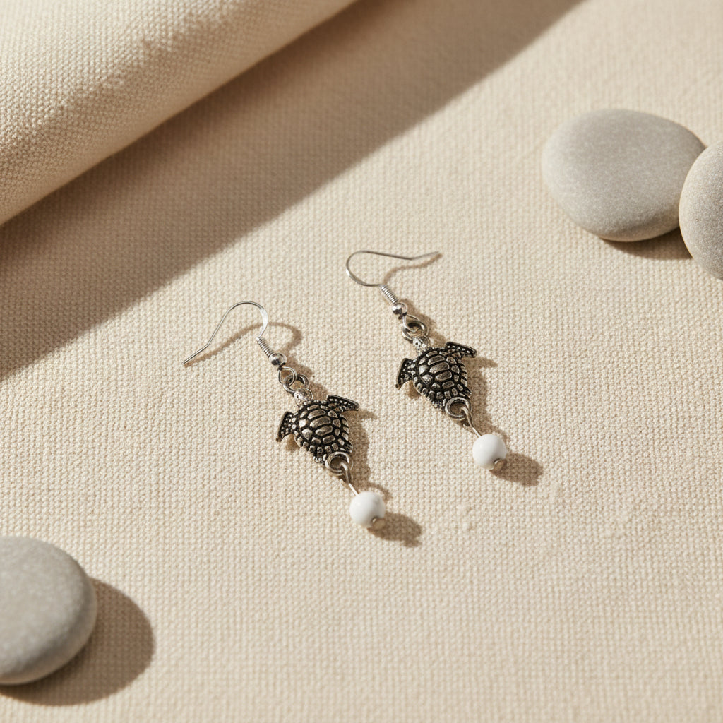 A pair of silver-toned turtle drop earrings with small white bead accents, displayed on a white card labeled River Inspired.