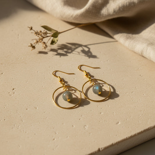 A pair of gold-tone dangle earrings with dark gray stone beads, displayed on a white card backing printed with the brand name 'River Inspired'.