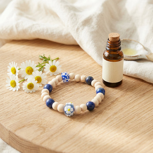 A beaded stretch bracelet featuring natural wood beads, dark blue glass beads, a blue and white patterned bead, and a small white and blue daisy charm, displayed on a plain light gray background.