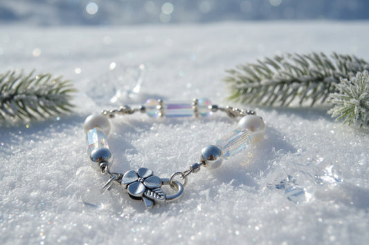 A silver chain bracelet featuring white faux pearls, iridescent clear beads, and a silver four-leaf clover charm with a dangling round bead, shown on a neutral gray background.