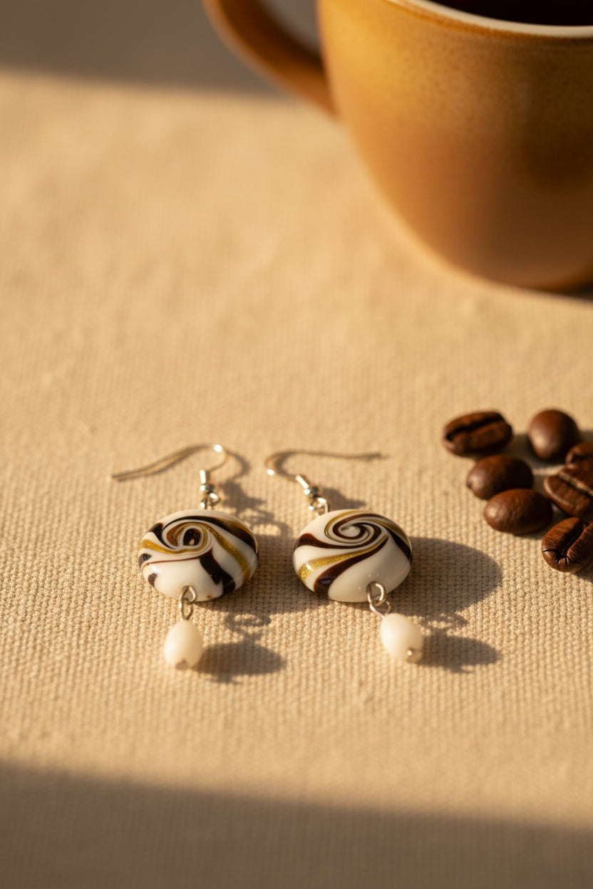 A pair of handmade dangle earrings displayed on a white card labeled 'River Inspired Handcrafted Jewelry.' The earrings have round marbled beads with black, white, and tan swirls, silver hooks, and small white bead drops, set against a plain light gray background.