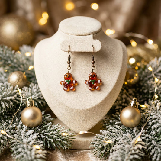 A pair of dangling earrings with small gingerbread man charms in orange-red glass, hanging from dark metal hooks against a plain light gray background.