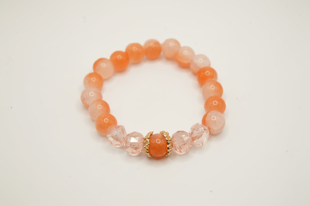 A beaded stretch bracelet featuring round orange and translucent pale pink beads, accented by faceted pink beads and a central round orange bead with gold-toned detailing, shown on a plain light gray background.