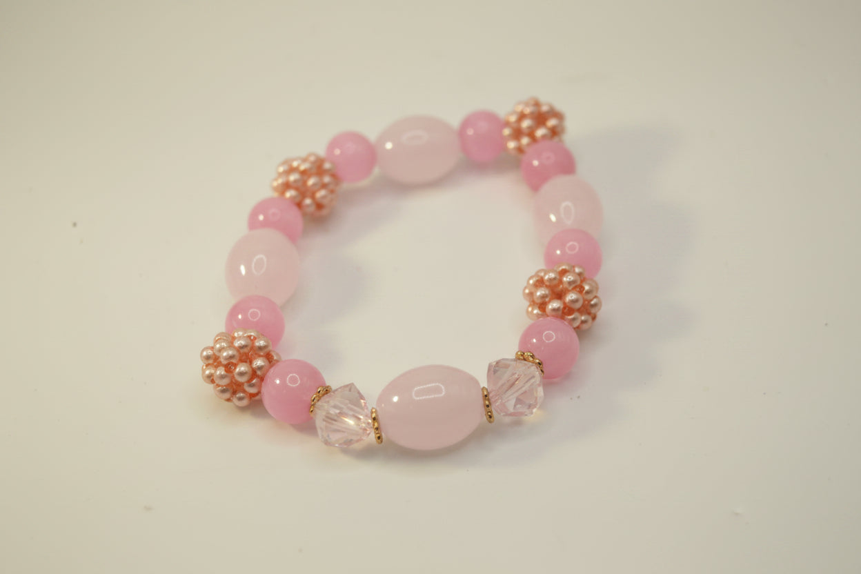 A pink beaded stretch bracelet featuring round pink beads, textured rose gold and pearl spacers, and two clear faceted crystal beads, displayed on a plain light gray background.