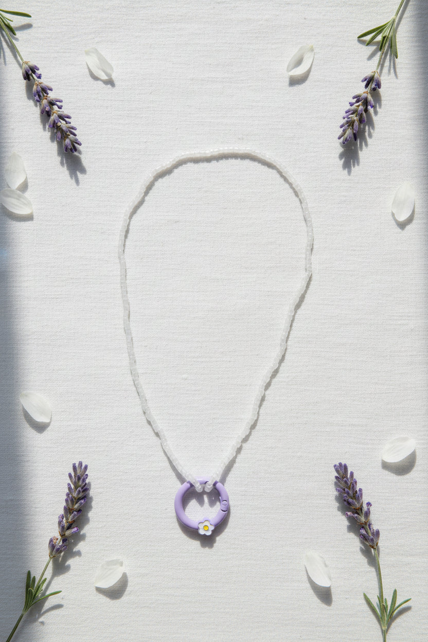A delicate silver-toned beaded necklace featuring a purple circular ring pendant with a small white flower detail at the bottom, displayed on a neutral gray background.