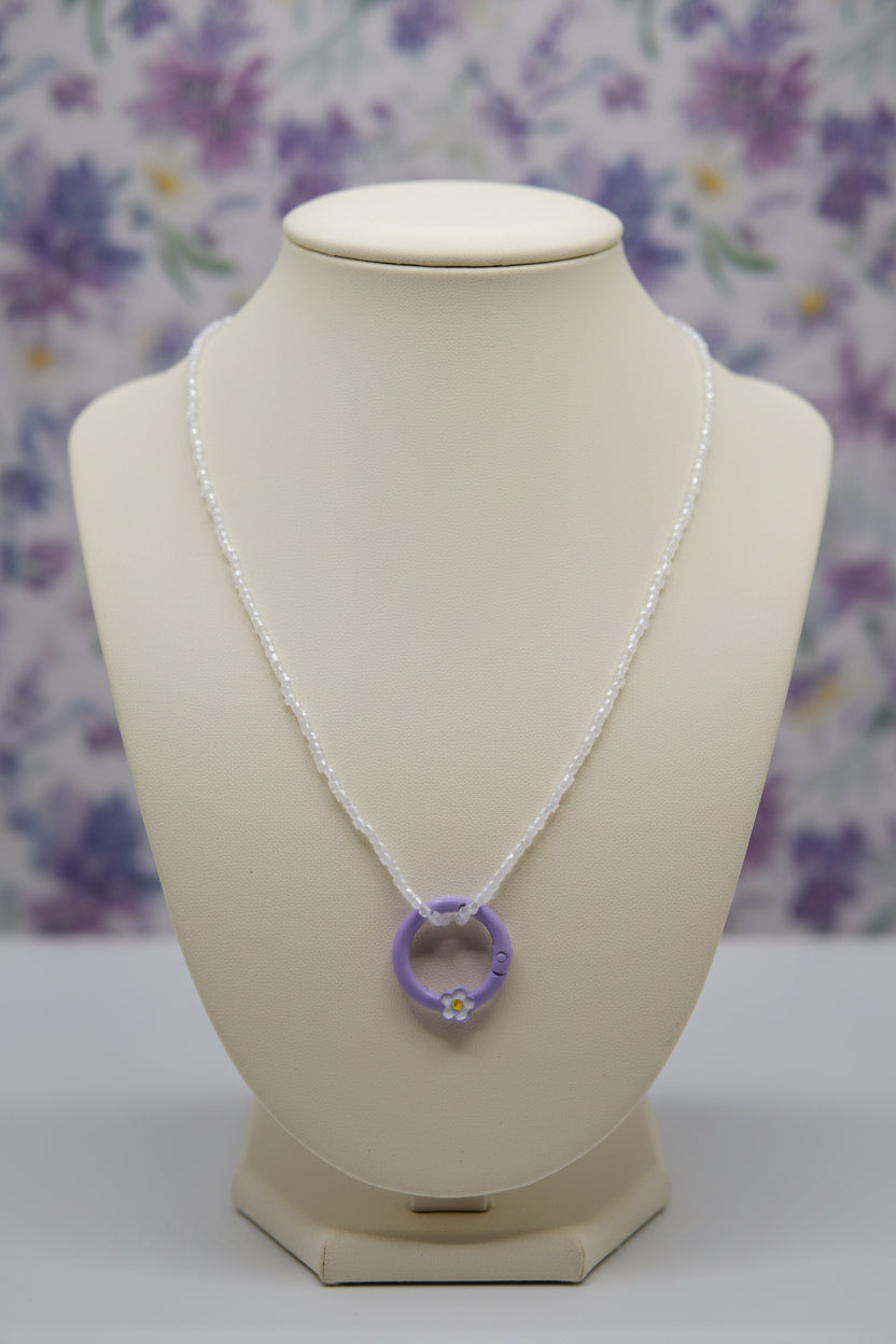 A delicate necklace featuring a small purple ring pendant with a tiny white flower charm, suspended from a thin white beaded chain, set against a plain light gray background.