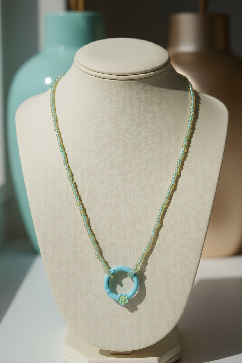 A delicate necklace featuring a small turquoise blue ring pendant with a green floral accent, suspended on a fine gold-toned chain against a plain light gray background.