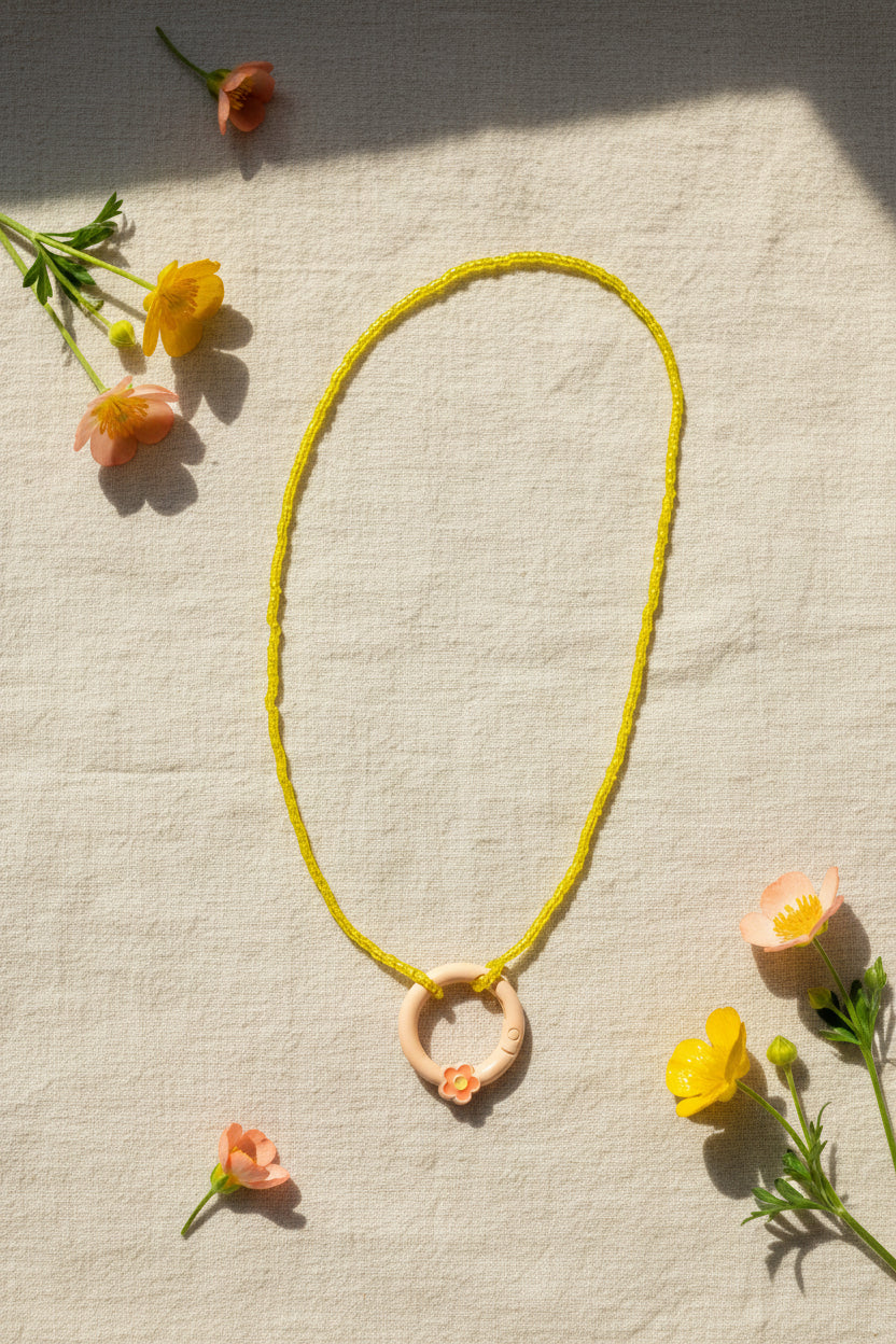 A yellow beaded necklace with a circular peach pendant featuring a small orange flower accent, displayed on a neutral gray background.